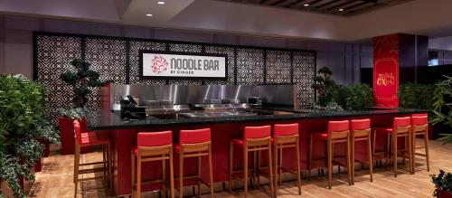 A modern noodle bar with a sleek counter, red seating, and decorative plants in a stylish interior setting.