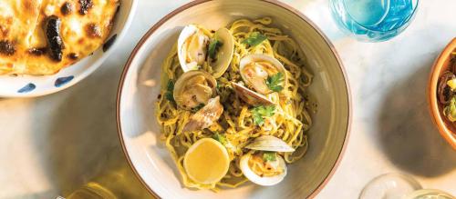 A bowl of yellow noodles topped with clams, lemon slices, and herbs, alongside a dish of baked cheese and a blue drink.