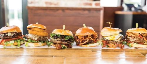 A lineup of six gourmet burgers, each with unique toppings, served on a wooden table in a casual dining setting.