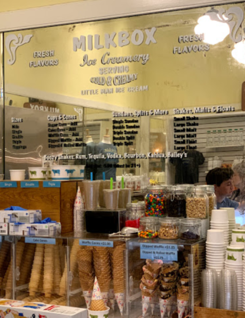 A menu board at Milkbox Ice Creamery displays flavors and prices, with ice cream cones and toppings in jars below.