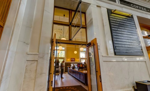 Entrance to a bright café with marble walls, featuring a menu board and wooden furniture.