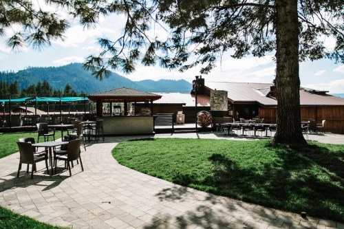 A scenic outdoor area with tables and chairs, surrounded by grass and trees, with mountains in the background.