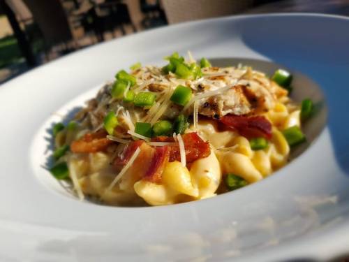 A close-up of creamy pasta topped with grilled chicken, bacon, green onions, and grated cheese in a white bowl.