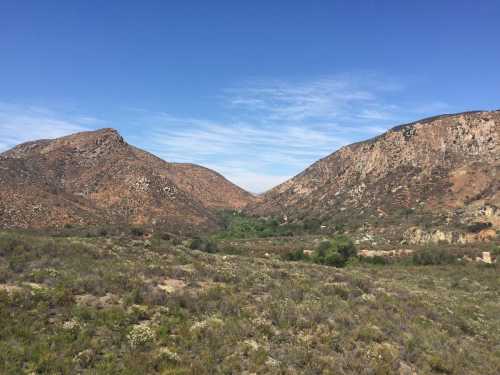 A scenic view of rugged mountains with a valley and blue sky, surrounded by green vegetation and rocky terrain.