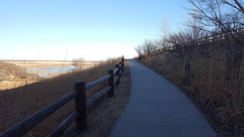 A paved path winds along a grassy hillside, bordered by a wooden fence and overlooking a calm body of water.