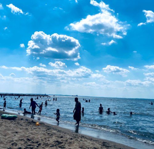 A sunny beach scene with people swimming and playing in the water under a blue sky with fluffy clouds.