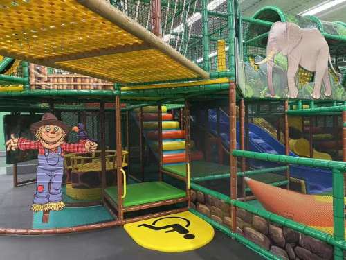 Colorful indoor playground with climbing structures, slides, and playful decorations, including a scarecrow and an elephant.