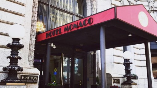 Entrance of Hotel Monaco with a red awning and decorative lamp posts, featuring the Kimpton logo.
