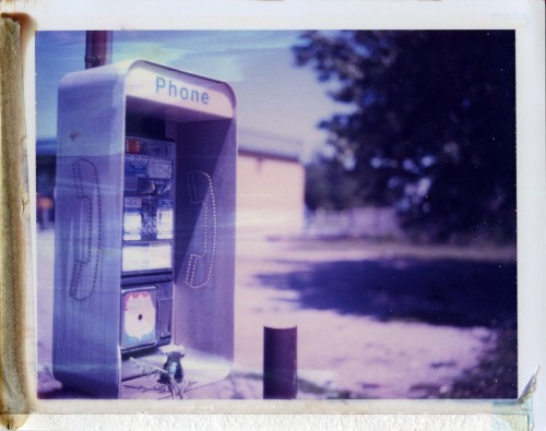 A vintage payphone stands alone in a sunny outdoor setting, surrounded by grass and trees.