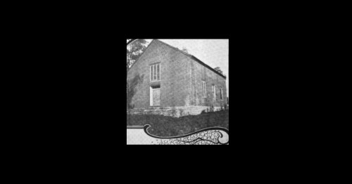 Black and white image of a brick building with a staircase and large windows, surrounded by greenery.