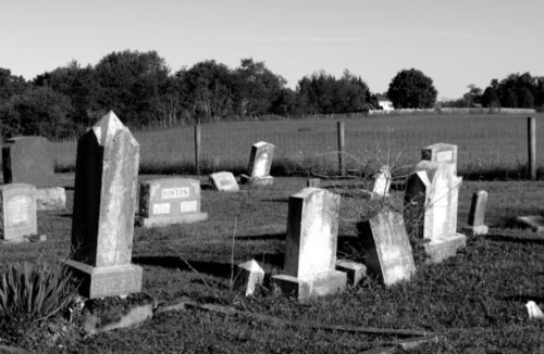 A black and white image of a cemetery with leaning gravestones and a grassy field in the background.