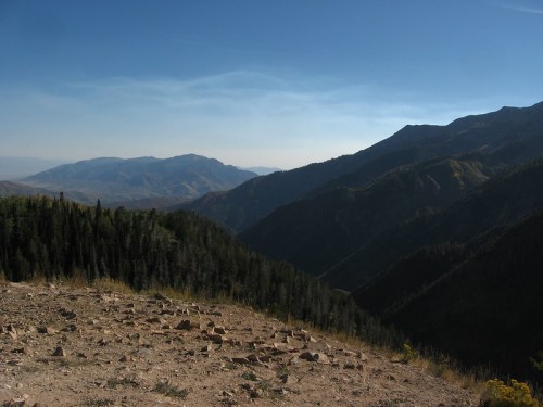 A panoramic view of mountains and valleys under a clear blue sky, with trees in the foreground and distant peaks.
