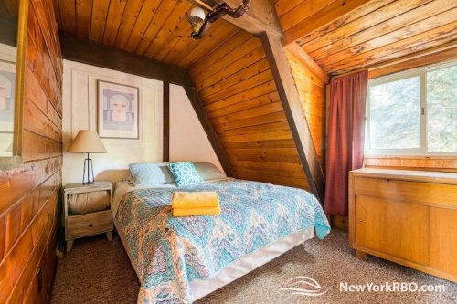 Cozy bedroom with wooden walls, a bed with a colorful quilt, a lamp, and a window with curtains.