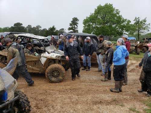 A group of people in rain gear gathers around muddy off-road vehicles in a wet, outdoor setting.
