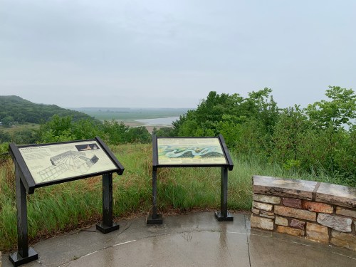 Two informational signs on a wet, grassy overlook with a view of a river and hills in the background on a cloudy day.