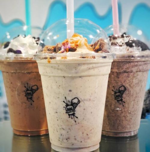 Three colorful milkshakes in clear cups, topped with whipped cream and various toppings, with a logo of a spider.