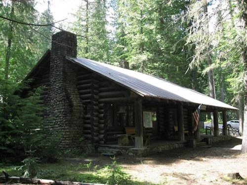 A rustic log cabin surrounded by tall trees, featuring a stone chimney and a sloped roof.