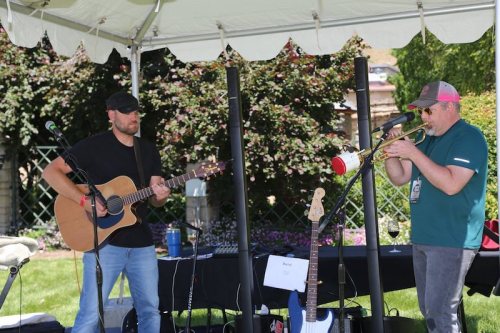 Two musicians perform under a tent; one plays guitar while the other plays trumpet, surrounded by greenery.