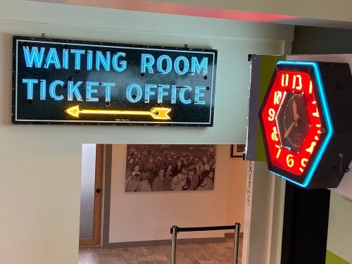 Neon sign for "Waiting Room Ticket Office" with an arrow, alongside a colorful wall clock.