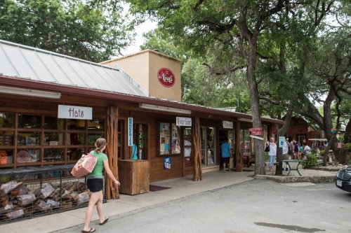 A woman walks past a rustic shop with wooden signage and trees, featuring various storefronts in a lively outdoor setting.