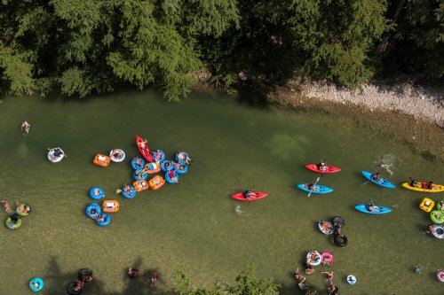 Aerial view of people kayaking and floating on colorful tubes in a clear river surrounded by greenery.