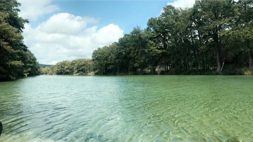 A serene lake surrounded by lush green trees under a partly cloudy sky. The water is clear and calm.