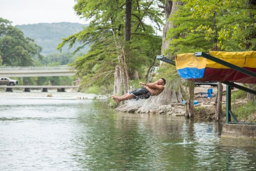 A person jumps off a dock into a river surrounded by trees, creating a splash in the water.