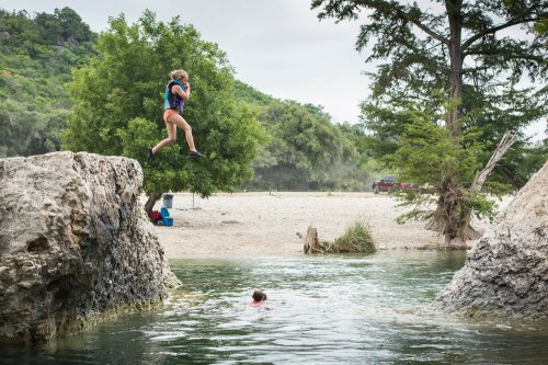 A person jumps off a rock into a swimming hole, while another person swims nearby in a scenic outdoor setting.