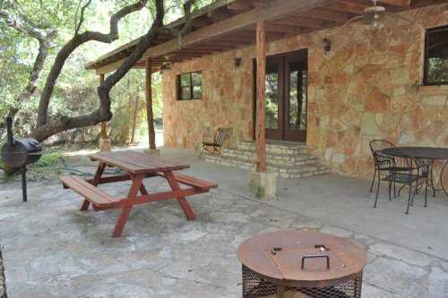 A rustic stone patio with a picnic table, chairs, and a fire pit, surrounded by trees and greenery.