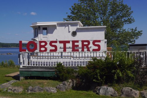 A white building with a large red sign reading "LOBSTERS," surrounded by greenery and a clear blue sky.