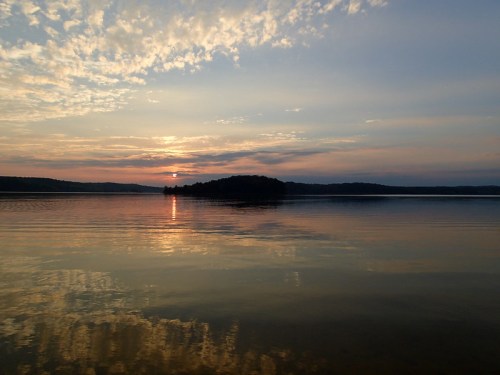 A serene lake at sunset, with soft clouds reflecting on the water and an island in the distance.