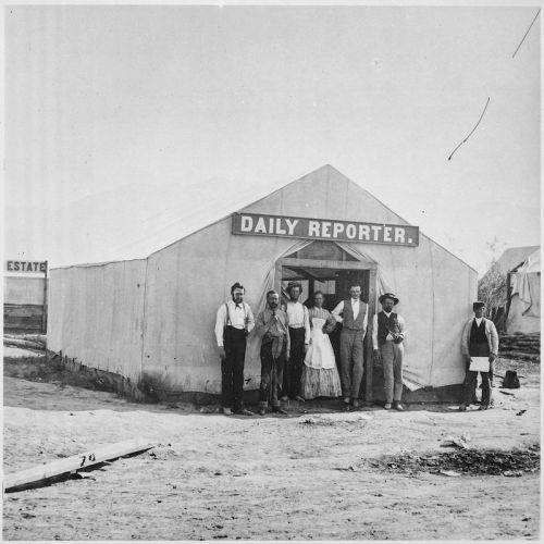 A group of people stands outside a tent labeled "Daily Reporter," with a dusty landscape in the background.