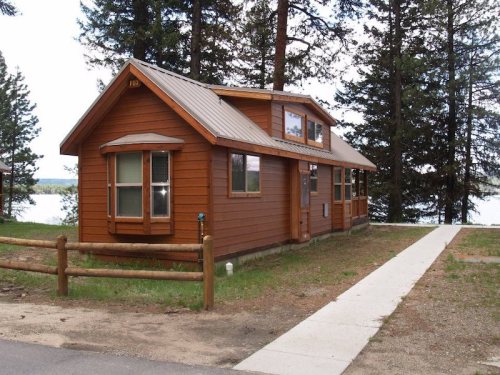 A cozy wooden cabin with a metal roof, surrounded by trees, near a lake and a pathway leading to the entrance.