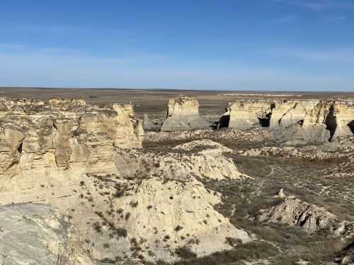 A vast landscape of rugged cliffs and rock formations under a clear blue sky, with sparse vegetation in the foreground.
