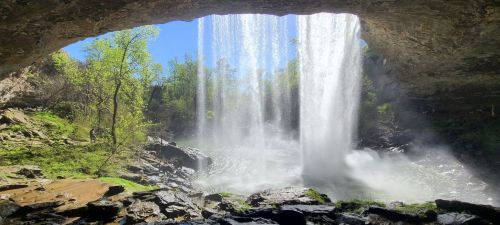 A stunning waterfall cascades over rocks, framed by a natural arch and surrounded by lush greenery and blue skies.
