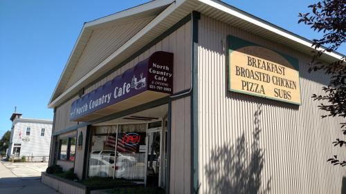Exterior of North Country Cafe, featuring signs for breakfast, broasted chicken, pizza, and subs. Open sign visible.