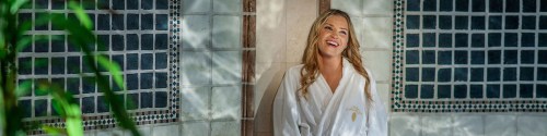 A smiling woman in a white robe stands in a spa-like setting with tiled walls and greenery.