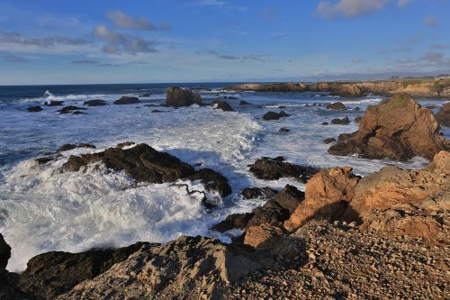 A rocky coastline with crashing waves under a blue sky and scattered clouds.