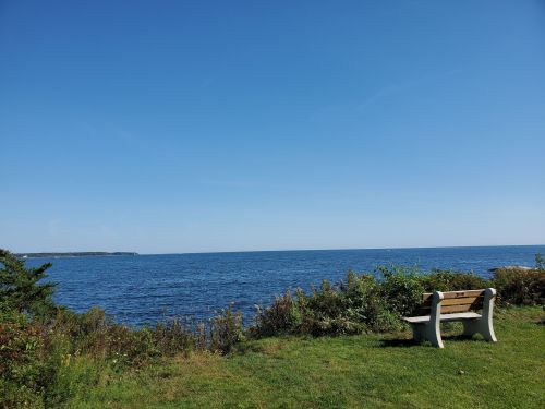 A serene coastal view featuring a bench overlooking a calm blue sea under a clear sky.