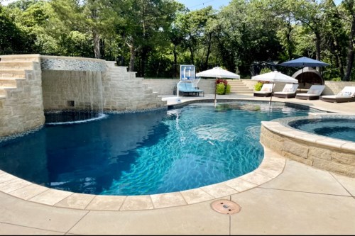 A luxurious outdoor pool with a waterfall, lounge chairs, umbrellas, and a hot tub surrounded by greenery.