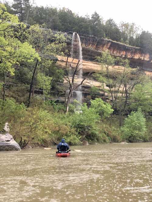 A person in a kayak paddles on a river, with a waterfall cascading down a rocky cliff in the background.