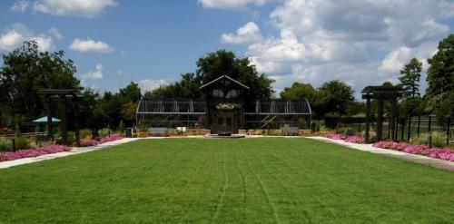 A lush green lawn leads to a garden building surrounded by colorful flowers and trees under a blue sky with clouds.
