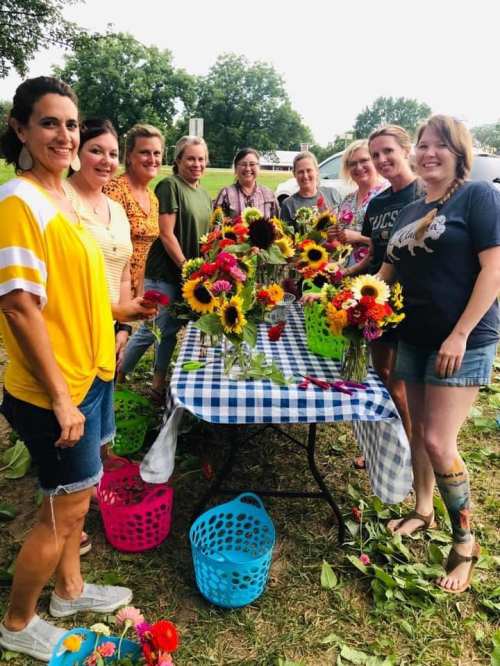 A group of women smiles around a table filled with colorful flower arrangements in a sunny outdoor setting.