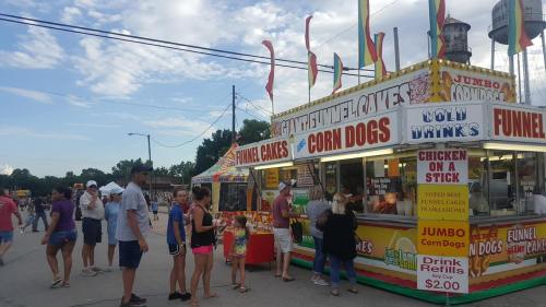 A colorful food stand at a fair, featuring funnel cakes and corn dogs, with people in line and a festive atmosphere.