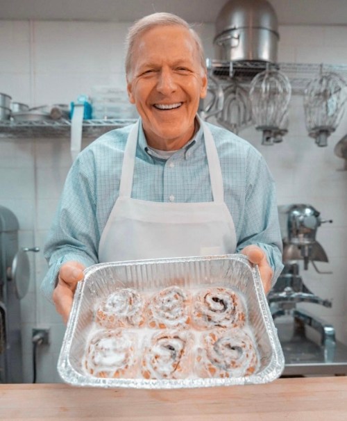 A smiling older man in an apron holds a tray of freshly baked cinnamon rolls in a kitchen setting.