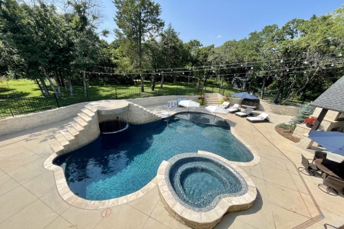 A spacious backyard with a pool shaped like a heart, a hot tub, and lounge chairs surrounded by greenery.