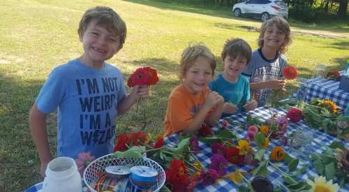 Four smiling children sit at a table covered in flowers, enjoying a fun activity outdoors.