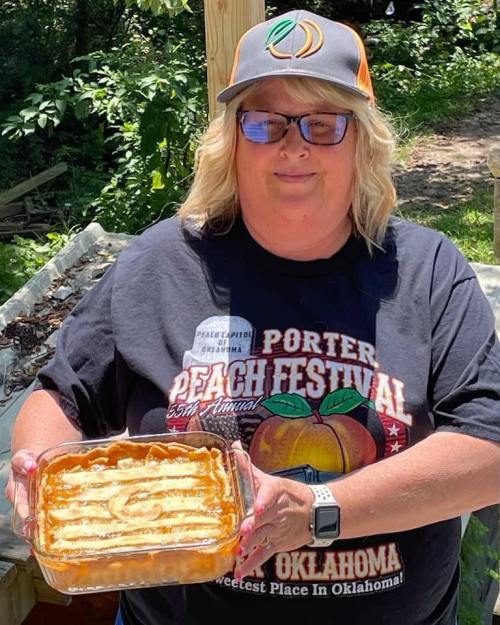 A smiling woman holds a dessert in a glass dish, wearing a cap and a t-shirt for the Porter Peach Festival.
