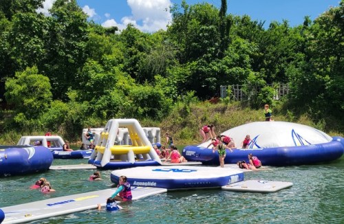 Children playing on inflatable water structures in a sunny outdoor setting surrounded by trees.