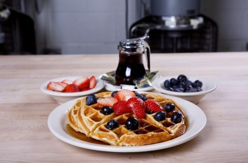 A plate of waffles topped with strawberries and blueberries, served with syrup and additional fruit on the side.